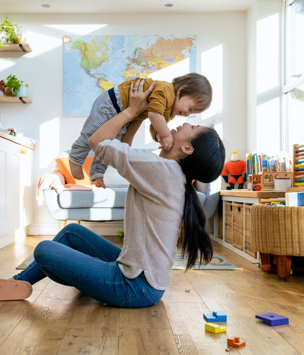 mid-adult mother sitting on the floor of a living room with her young boy she is lifting him up in the air